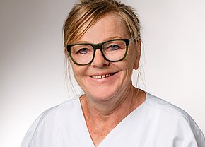 Station 10 Eine lächelnde Frau mit blonden Haaren trägt eine weiße Arbeitskleidung und grüne Brille. Ihr Gesichtsausdruck vermittelt Freundlichkeit und Professionalität, was auf einen Gesundheitsberuf hinweisen könnte. Der Hintergrund ist neutral und unauffällig, um den Fokus auf die Person zu lenken.