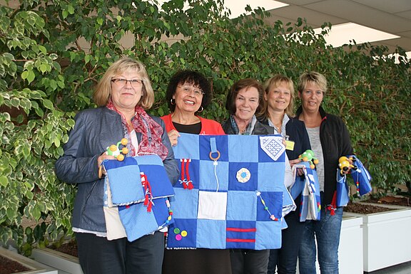 Fünf Frauen stehen zusammen und halten mehrere blau-weiße Fühl- bzw. Beschäftigungsdecken mit Knöpfen, Reißverschlüssen und Bändern in die Kamera. Sie lächeln; es wirkt wie eine Präsentation selbstgemachter Therapie- oder Demenzdecken.