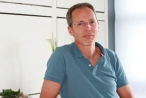 Dietrich de Fallois Ein Mann mit brünettem, kurzem Haar und einer Brille sitzt mit einem nachdenklichen Ausdruck. Er trägt ein beige-blaues Polohemd. Im Hintergrund sind Pflanzen sichtbar. Die Umgebung wirkt hell und freundlich.