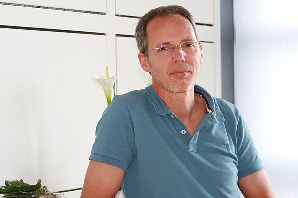 Ein Mann mit brünettem, kurzem Haar und einer Brille sitzt mit einem nachdenklichen Ausdruck. Er trägt ein beige-blaues Polohemd. Im Hintergrund sind Pflanzen sichtbar. Die Umgebung wirkt hell und freundlich.