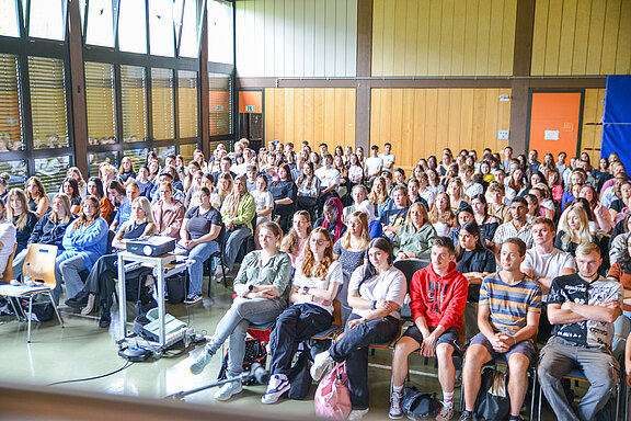 Ein großes Publikum sitzt in einem Raum mit Holzverkleidung, aufmerksam auf eine Präsentation ausgerichtet. Die Zuschauer sind vorwiegend Jugendliche und junge Erwachsene, die in ihren Sitzen stehen und auf den Bildschirm focusiert sind.
