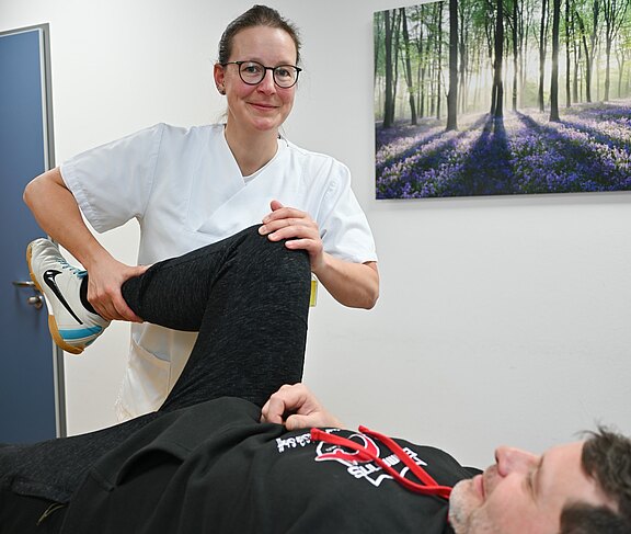 Physiotherapeutin behandelt einen Patienten: Der Mann liegt auf einer Liege, sie beugt sein rechtes Bein und hält das Knie, um eine Mobilisations- beziehungsweise Dehnübung des Beins und Hüftgelenks durchzuführen.