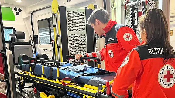 Zwei Rettungskräfte des Deutschen Roten Kreuzes laden eine Krankentrage in einen Rettungswagen. Sie prüfen Ausrüstung und sichern die Trage auf der ausziehbaren Plattform. Szene zeigt Vorbereitung für einen Notfalleinsatz im Krankenwagen.