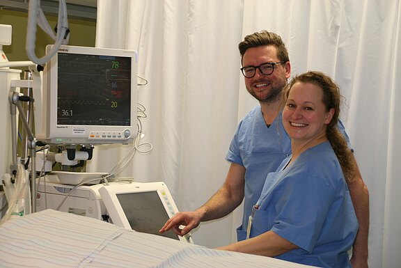 Zwei medizinische Fachkräfte in blauer OP-Kleidung stehen neben einem Patientenmonitor. Sie bedienen das Gerät und kontrollieren Vitaldaten auf dem Bildschirm. Die Szene wirkt wie in einem Krankenhausraum mit Vorhängen; beide lächeln.
