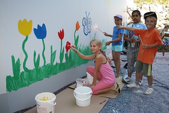 Mehrere Kinder malen gemeinsam ein Wandbild: bunte Blumen, Gras und ein Schmetterling auf einer hellen Mauer. Daneben stehen Farbeimer und Pinsel. Gemeinschaftliche Malaktion im Freien.