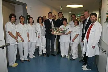 Eine Gruppe von Pflegekräften und Ärzten steht in einem Krankenhausflur. In der Mitte hält eine Person einen großen Blumenstrauß. Alle lächeln und wirken freundlich. Die Atmosphäre ist festlich und herzlich.