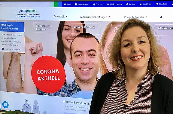 Eine Frau lächelt vor einem Hintergrund, der eine Website des Klinikum Bayreuth zeigt. Auf der Website ist ein rotes Feld mit der Aufschrift „CORONA AKTUELL“ und Bilder von Personen im medizinischen Kontext zu sehen.