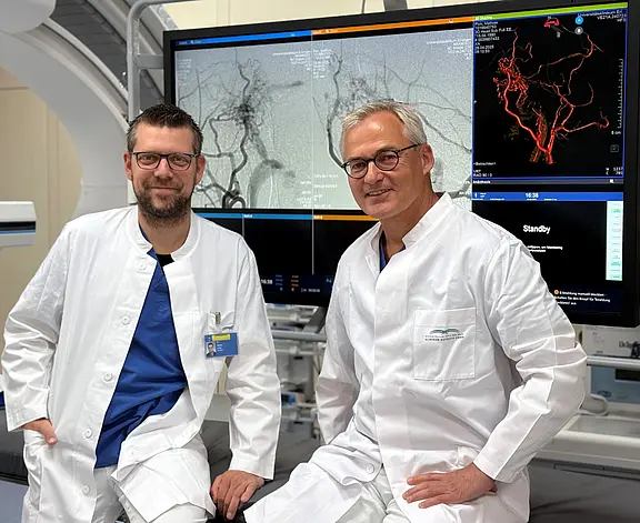 Zwei Ärzte in weißen Kitteln sitzen in einem Klinikraum vor großen Monitoren. Auf den Bildschirmen sind Angiografie-Aufnahmen von Gehirn- und Halsgefäßen zu sehen. Die Szene zeigt eine radiologische beziehungsweise neuroangiografische Arbeitsumgebung.