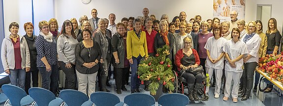 Große Gruppe von Mitarbeitenden posiert in einem hellen Raum für ein Gruppenfoto. Einige tragen weiße Pflegekleidung. In der Mitte festliche Weihnachtsdeko mit Tannenzweigen und roten Kugeln. Stimmung: gemeinsames Team- oder Feierfoto.
