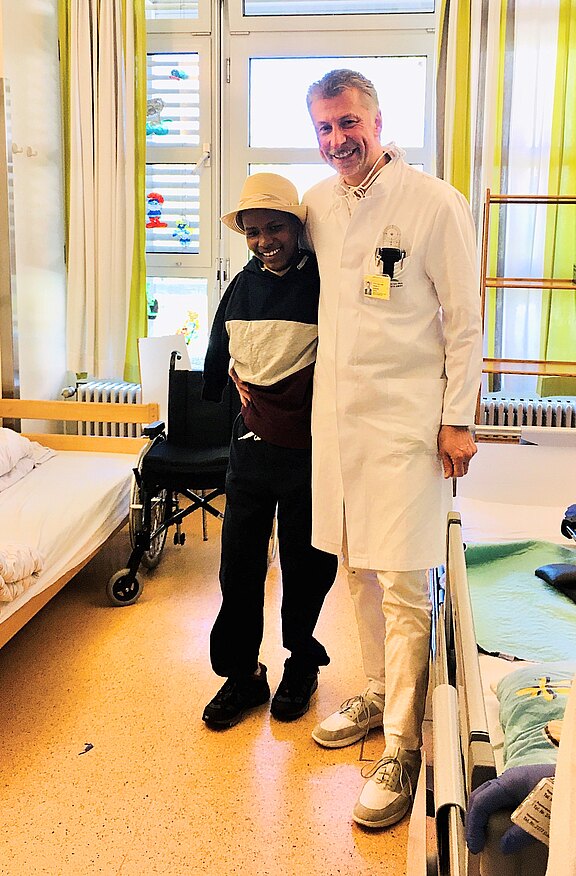 Krankenhauszimmer: Eine Person im weißen Kittel lächelt und stützt eine andere Person; neben ihnen stehen Bett und Rollstuhl.