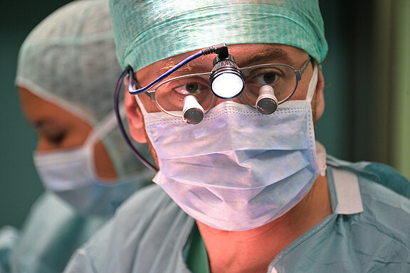 Ein Chirurg mit einer grünen OP-Kappe und einer Schutzmaske sieht konzentriert aus. Er trägt eine Brille mit integrierter Beleuchtung. Im Hintergrund ist eine andere Person in OP-Kleidung zu erkennen. Die Szene vermittelt den Eindruck einer chirurgischen Umgebung.