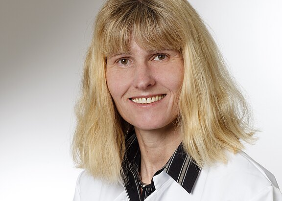 Eine lächelnde Frau mit langen, blonden Haaren trägt einen weißen Kittel und ein schwarz-weiß gestreiftes Hemd. Der Hintergrund ist hell und neutral gehalten.