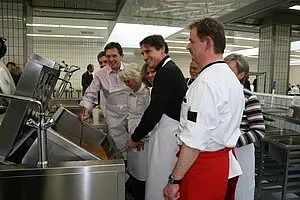 Eine Gruppe von Personen in Kochschürzen steht um einen großen Kochtopf in einer Küche. Einige Menschen rühren in der Mischung, während andere zuschauen und lachen. Die Atmosphäre ist freundlich und einladend, mit einer gemeinsamen Begeisterung für das Kochen.