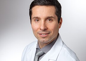 Porträt eines erwachsenen Mannes in weißem Kittel, grauem Hemd und gestreifter Krawatte. Er blickt freundlich in die Kamera vor neutralem, hellgrauem Hintergrund.