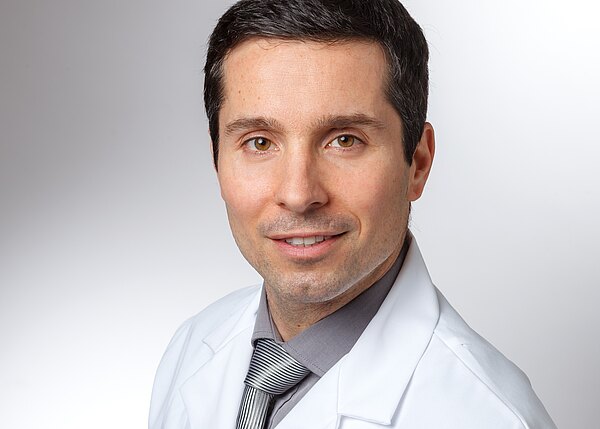 Porträt eines erwachsenen Mannes in weißem Kittel, grauem Hemd und gestreifter Krawatte. Er blickt freundlich in die Kamera vor neutralem, hellgrauem Hintergrund.