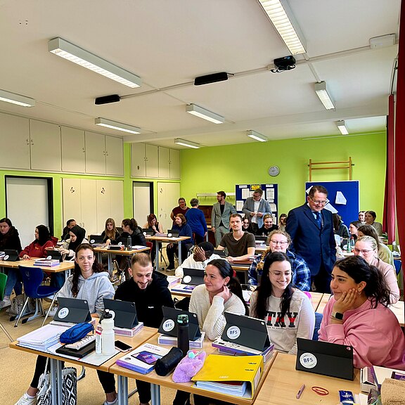 Lebendiges Klassenzimmer: Viele junge Erwachsene sitzen an Gruppentischen, arbeiten mit Tablets und Notizen und sprechen miteinander. Ein Lehrender geht durch den Raum und unterstützt Lernende. Die Stimmung wirkt aufmerksam und freundlich, Fokus auf gemeinsames Lernen mit digitalen Geräten.