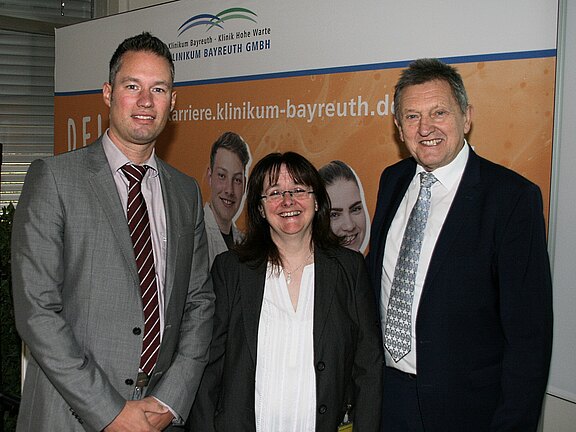 Drei Menschen in Businesskleidung – zwei Männer und eine Frau – stehen nebeneinander und lächeln in die Kamera. Im Hintergrund ein Banner des Klinikums Bayreuth, Anlass wirkt offiziell.