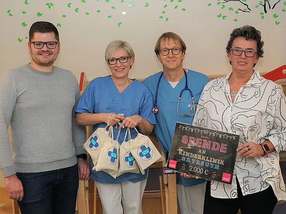 Vier Erwachsene stehen zusammen in einer Klinik. Zwei tragen OP‑Kleidung, einer mit Stethoskop. Eine Person hält mehrere kleine Geschenkbeutel, eine andere ein Schild: „Spende an Kinderklinik Bayreuth – 2.000 €“. Die Gruppe lächelt und präsentiert die Spendenübergabe.