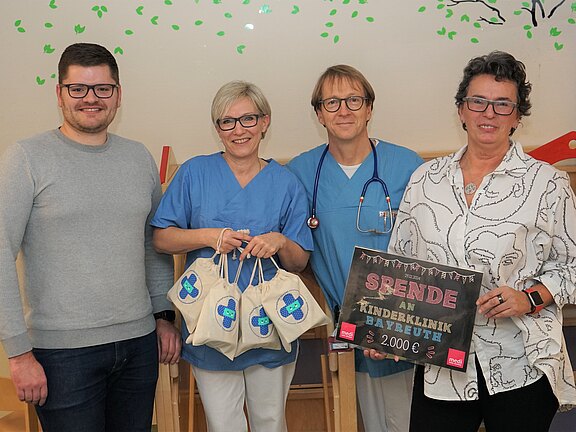 Vier Erwachsene stehen zusammen in einer Klinik. Zwei tragen OP‑Kleidung, einer mit Stethoskop. Eine Person hält mehrere kleine Geschenkbeutel, eine andere ein Schild: „Spende an Kinderklinik Bayreuth – 2.000 €“. Die Gruppe lächelt und präsentiert die Spendenübergabe.