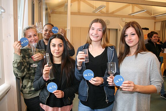 Fünf Jugendliche in einem Klassenraum halten Trinkgläser und blaue runde Karten mit Aufdrucken wie „ca. 12 g“ und „ca. 1,9 g“. Die Szene wirkt wie eine Aufklärung über Alkoholmengen.