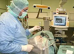 Ein Chirurg in Schutzkleidung führt einen Eingriff durch. Auf einem Monitor im Hintergrund sind medizinische Bilder zu sehen. Der Patient liegt auf dem Bett, teilweise abgedeckt, während der Arzt mit chirurgischen Instrumenten arbeitet.