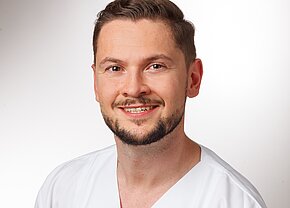 Station 55 Ein lächelnder Mann mit kurzen, braunen Haaren und einem Bart trägt ein weißes medizinisches Outfit. Er hat einen freundlichen Gesichtsausdruck und steht vor einem hellen, einfarbigen Hintergrund.