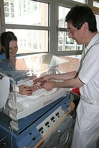 Eine Frau und ein Mann stehen an einem Inkubator in einem Krankenhaus. Der Mann, der in weißer Kleidung ist, kümmert sich um ein Neugeborenes, während die Frau ihm dabei hilft. Das Bild vermittelt eine fürsorgliche Atmosphäre und zeigt die medizinische Betreuung eines Säuglings.