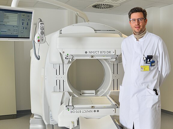 Medizinischer Mitarbeiter in weißem Kittel steht neben einem offenen CT-/Nuklearmedizin-Scanner mit ringförmiger Öffnung und Patiententisch. Ein Monitor und Bedienelemente sind sichtbar. Klinischer Untersuchungsraum, bereit für eine bildgebende Untersuchung.