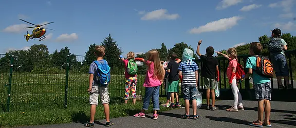 Mehrere Kinder mit Rucksäcken stehen am Zaun und schauen einem gelben Hubschrauber zu, der knapp über einer Wiese fliegt oder landet. Sonniger Himmel.
