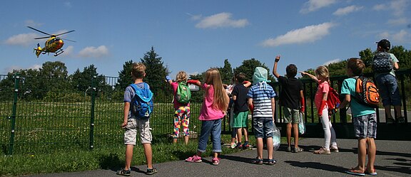 Mehrere Kinder mit Rucksäcken stehen am Zaun und schauen einem gelben Hubschrauber zu, der knapp über einer Wiese fliegt oder landet. Sonniger Himmel.