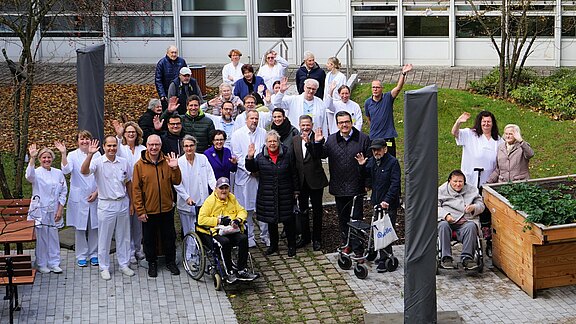 Große Gruppe aus Pflegepersonal und Bewohnern/Patienten vor einem Gebäude; einige mit Rollstuhl oder Rollator. Alle posieren im Hof, lächeln und winken in die Kamera, wie bei einem Gruppenfoto einer Pflegeeinrichtung.