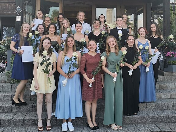 Gruppe junger Menschen in festlicher Kleidung posiert auf einer Treppe. Viele halten weiße Rosen und einige Dokumente in der Hand. Alle lächeln in die Kamera, als ob sie einen Abschluss oder eine Feier begehen.