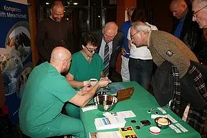 Zwei Personen in grüner medizinischer Kleidung demonstrieren einem interessierten Publikum an einem Tisch. Um sie herum stehen mehrere ältere Männer, die aufmerksam zuschauen. Auf dem Tisch liegen medizinische Utensilien und Informationsmaterialien.