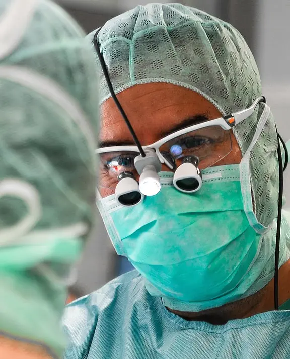 Chirurg im Operationssaal, mit grüner OP‑Kleidung, Haube, Maske und Vergrößerungsbrille, konzentriert auf einen Eingriff; im Vordergrund verschwommen eine weitere OP-Person.