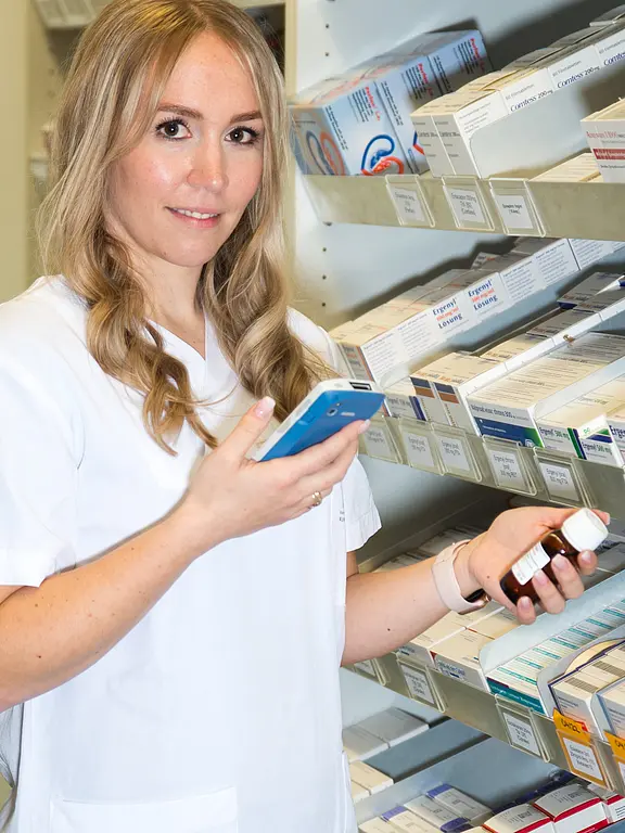 Eine Frau in einem weißen Kittel steht vor einem Regalsystem voller Arzneimittel. Sie hält ein Rezeptbuch in der linken Hand und ein Handy in der rechten Hand, während sie auf das Handy schaut. Ihr Gesicht zeigt Konzentration und Engagement.