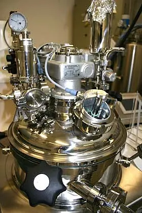 Ein hochglänzender, runder Druckbehälter aus Edelstahl mit verschiedenen Ventilen, Druckmanometern und Rohren. Die komplexe Anordnung von mechanischen Teilen deutet auf eine technische Anwendung hin, möglicherweise in einem Labor oder industriellen Umfeld.