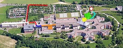 Luftaufnahme eines Geländes mit mehreren Gebäuden und Grünflächen. Farbcodierte Wege (rot, grün, blau, orange) zeigen verschiedene Routen zu einem markierten Punkt. Die Umgebung wirkt weitläufig und strukturiert, mit Parkplätzen und weiteren Einrichtungen im Hintergrund.