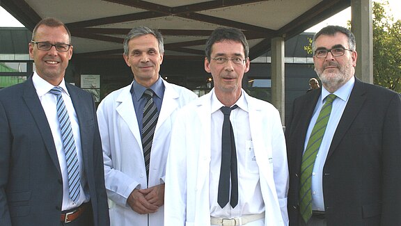 Vier Männer stehen vor einem modernen Gebäude. Drei von ihnen tragen weiße Laborkittel, während einer ein Anzugshirt und eine Krawatte trägt. Die Gruppe wirkt freundlich und professionell. Im Hintergrund sind unscharfe Details der Umgebung sichtbar.