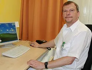 Ein Mann sitzt an einem Schreibtisch vor einem Computer. Er trägt ein helles Hemd und hat einen freundlichen Gesichtsausdruck. Im Hintergrund sind eine orangefarbene Vorhänge und ein Monitor mit einer Desktop-Oberfläche sichtbar.