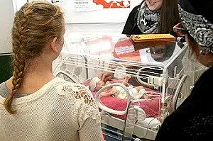 Eine Frau mit geflochtenem Haar steht vor einem Inkubator, in dem ein Neugeborenes liegt. Im Hintergrund sprechen zwei weitere Frauen miteinander. Die Szene vermittelt den Moment der Beobachtung und des Austausches über das Baby im Inkubator.