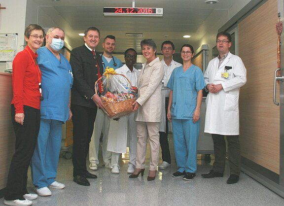 In einem Krankenhausflur steht eine Gruppe von Ärzten und Pflegekräften. Zwei Besucher überreichen gemeinsam einen großen Geschenkkorb an das Team. Alle lächeln in die Kamera; es wirkt wie eine feierliche Dankesgeste, vermutlich zu Weihnachten.