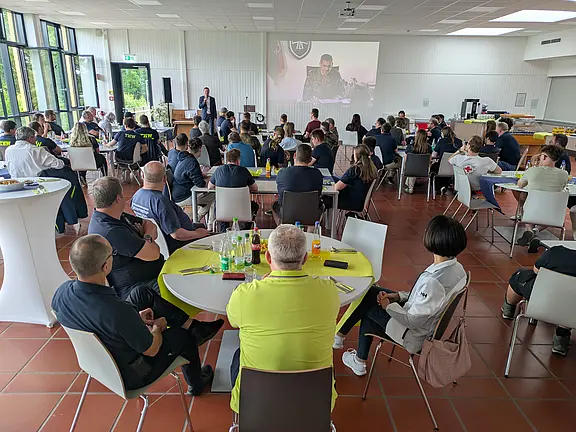Eine Gruppe von Menschen sitzt in einem großen Raum mit Tischen und Stühlen, während ein Mann auf einer Bühne spricht. Im Hintergrund ist eine Präsentation zu sehen. Die Teilnehmer tragen einheitliche T-Shirts und hören aufmerksam zu. Es herrscht eine freundliche, engagierte Atmosphäre.
