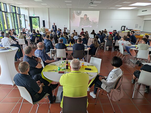 Eine Gruppe von Menschen sitzt in einem großen Raum mit Tischen und Stühlen, während ein Mann auf einer Bühne spricht. Im Hintergrund ist eine Präsentation zu sehen. Die Teilnehmer tragen einheitliche T-Shirts und hören aufmerksam zu. Es herrscht eine freundliche, engagierte Atmosphäre.
