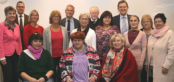 Gruppenfoto: Fünfzehn Erwachsene posieren in einem hellen Raum. Drei Personen sitzen vorne, die anderen stehen dahinter. Unterschiedliche Kleidung und Alter, freundliche Gesichter. Es wirkt wie ein Treffen oder eine Feier in einer Institution oder einem Verein.