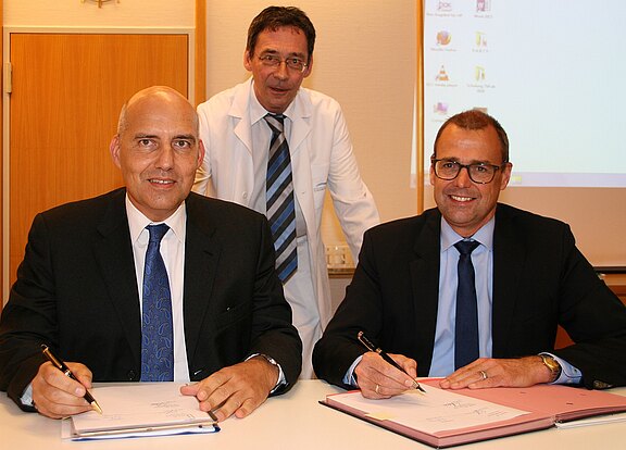 Zwei Männer im Anzug sitzen an einem Tisch und unterschreiben Dokumente. Hinter ihnen steht ein Mann im weißen Kittel und beobachtet. Formelle Szene, vermutlich Vertragsunterzeichnung oder Kooperation im medizinischen Umfeld.