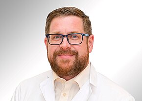 Ein Mann mit Brille und einem Bart lächelt freundlich in die Kamera. Er trägt einen weißen Laborkittel und ein helles Hemd. Der Hintergrund ist sanft gradientiert von Grau zu Weiß.