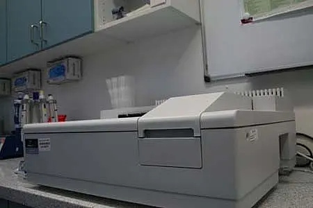 Ein Laborgerät auf einer Tischfläche. Es handelt sich um einen grauen Analyse- oder Messapparat mit einer offenen Ladefläche, um Proben einzuführen. Im Hintergrund sind einige medizinische Utensilien und ein Regal zu sehen, die jedoch nicht im Fokus stehen.