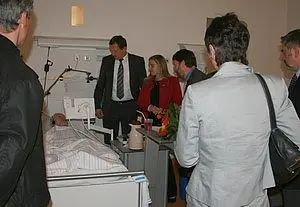 Eine Gruppe von sechs Personen steht um ein Krankenhausbett. Eine Person im Bett ist sichtbar. Die Anwesenden scheinen sich um den Patienten zu kümmern oder ihn zu besuchen. Die Atmosphäre wirkt ernst und fürsorglich.