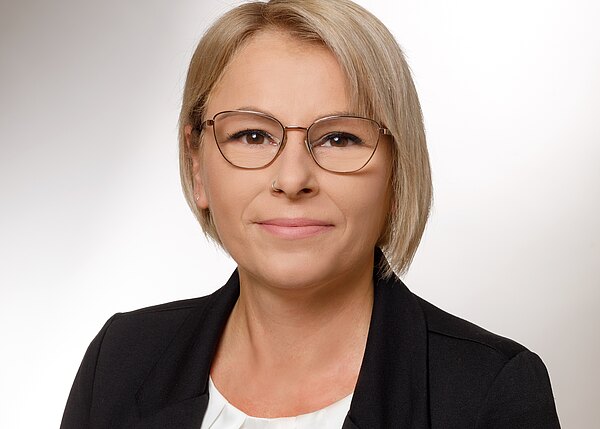 Porträtfoto einer erwachsenen Person mit kurzen blonden Haaren und runder Brille. Die Person trägt schwarzen Blazer über weißer Bluse, blickt direkt in die Kamera; neutraler Hintergrund, ruhiger, professioneller Ausdruck.