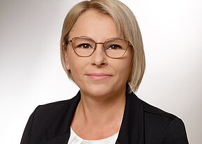 Nicole Embacher Porträtfoto einer erwachsenen Person mit kurzen blonden Haaren und runder Brille. Die Person trägt schwarzen Blazer über weißer Bluse, blickt direkt in die Kamera; neutraler Hintergrund, ruhiger, professioneller Ausdruck.